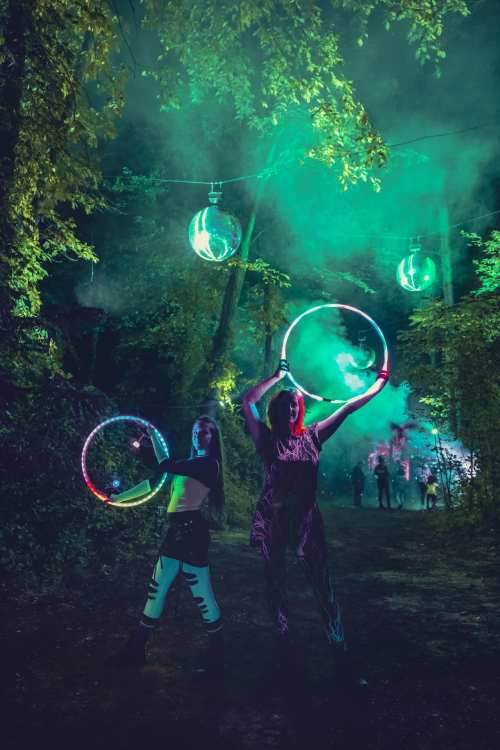 Two performers with glowing hula hoops pose in a misty forest, surrounded by colorful lights and disco balls.