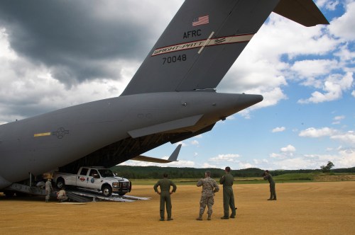 Military personnel unload a truck from the rear of a large cargo aircraft on a grassy field under a cloudy sky.