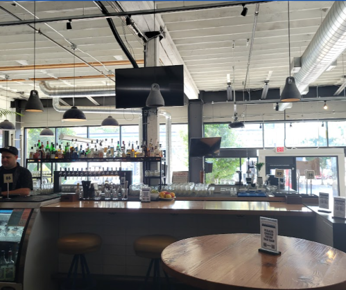 A modern bar interior with a bartender, shelves of liquor, and large windows letting in natural light.