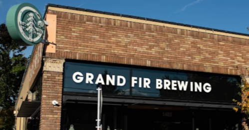 Exterior of Grand Fir Brewing, a brick building with large windows and a sign, surrounded by trees and a clear blue sky.