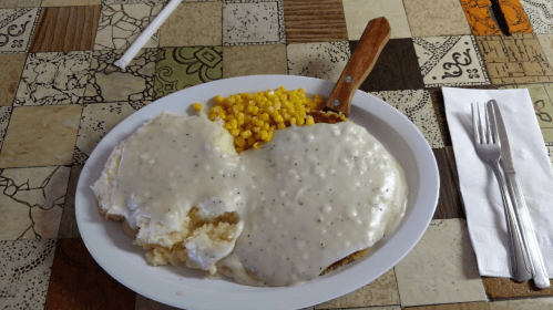 A plate of chicken fried steak smothered in gravy, served with mashed potatoes and corn on a patterned table.