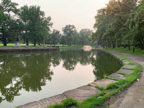 A serene park scene featuring a calm pond, lush trees, and a fountain, with a pathway along the water's edge.