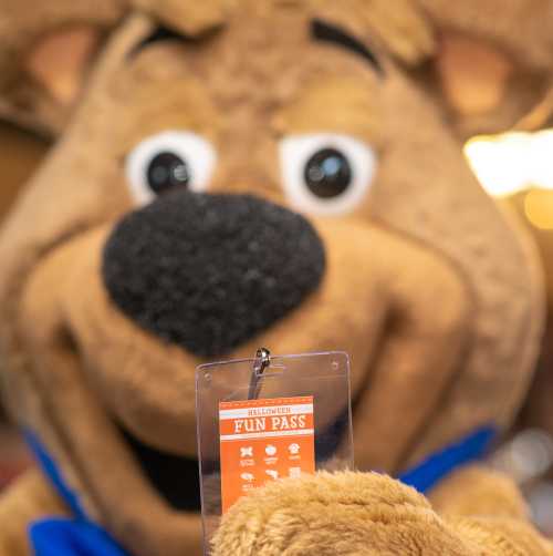 A cartoon dog mascot holds up a Halloween Fun Pass, smiling at the camera.