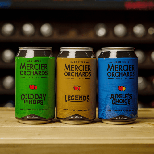 Three cans of Mercier Orchards hard cider: "Cold Day in Hops," "Legends," and "Adel's Choice," displayed on a wooden surface.