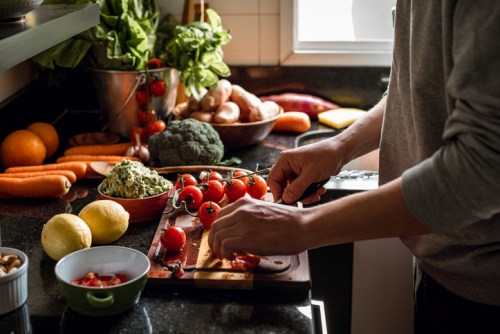 A person chops tomatoes on a cutting board surrounded by fresh vegetables and fruits in a bright kitchen.