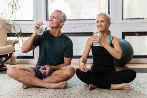 A man and woman sit cross-legged on a mat, smiling and drinking water after a workout in a bright, airy room.