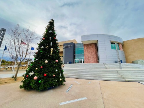 A large Christmas tree adorned with ornaments stands in front of Henderson City Hall, with a cloudy sky above.