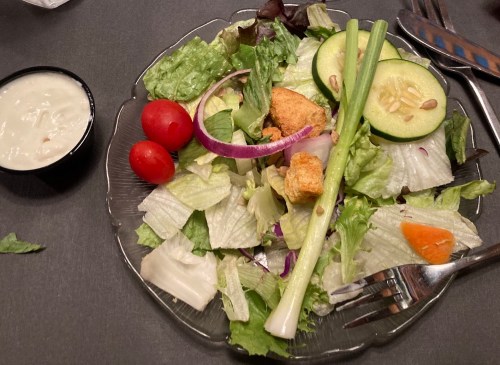A fresh salad with lettuce, cucumbers, cherry tomatoes, red onion, and a side of dressing on a dark table.