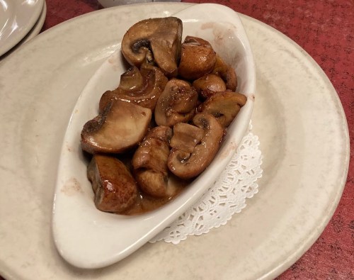 A white dish filled with cooked mushrooms in a savory sauce, placed on a red tablecloth.