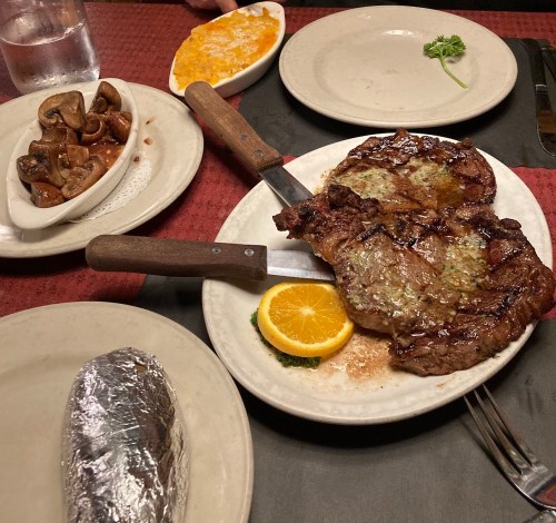 A plate with a large steak topped with herb butter, an orange slice, a baked potato, and sides of mushrooms and cheese.