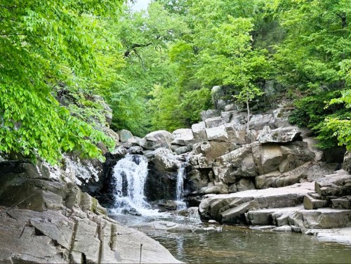 A serene waterfall cascades over rocks, surrounded by lush green trees and a calm pool of water.