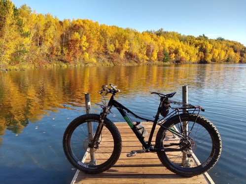 A black fat bike on a wooden dock by a calm lake, surrounded by vibrant autumn foliage.