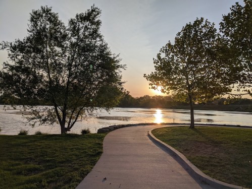 A winding path by a lake at sunset, framed by trees and reflecting warm light on the water's surface.