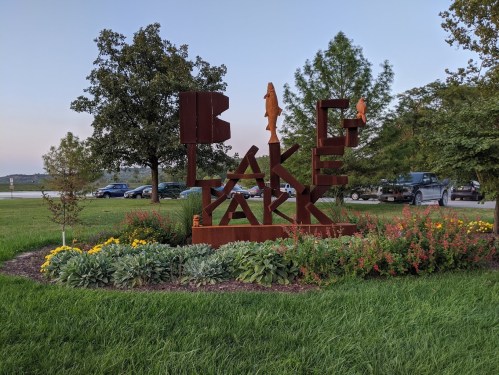 Sign for Lake Park with metal letters and fish sculptures, surrounded by colorful flowers and greenery.