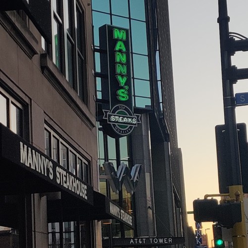 Neon sign for Manny's Steakhouse on a city street, with the AT&T Tower in the background at sunset.