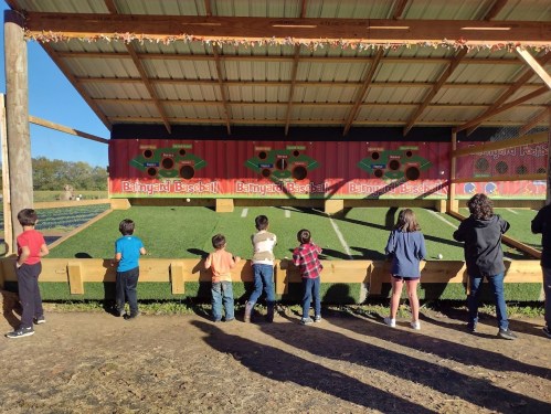 Children watch a baseball-themed attraction with colorful decorations and a grassy area for play.