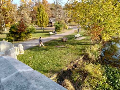 A park scene with trees, benches, and a person walking near a pond on a sunny day.