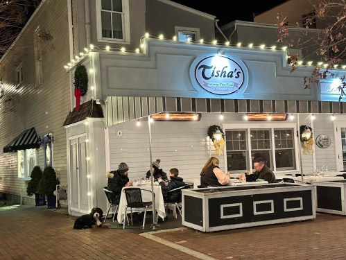 A cozy restaurant exterior with festive lights, featuring diners at tables and a dog resting nearby.