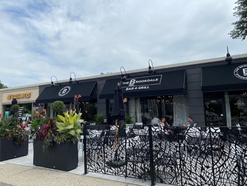Exterior view of The Brookdale Bar + Grill, featuring outdoor seating and decorative plants, under a cloudy sky.