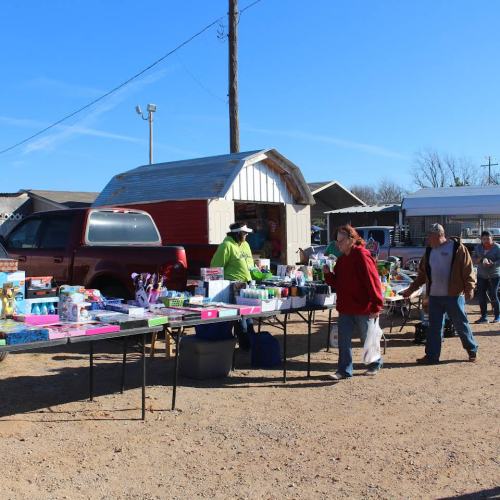 A bustling outdoor market with tables of various goods, people browsing, and a red truck in the background under a clear blue sky.