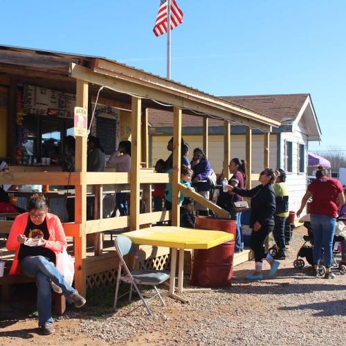 A busy outdoor food stand with people eating, ordering, and enjoying the sunny day, with an American flag in the background.