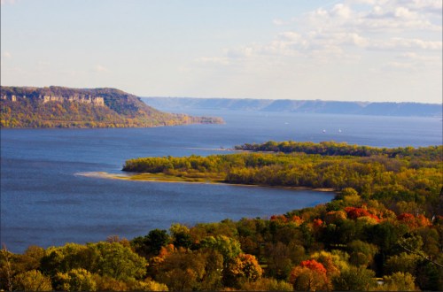 A scenic view of a river surrounded by lush greenery and autumn-colored trees under a blue sky.
