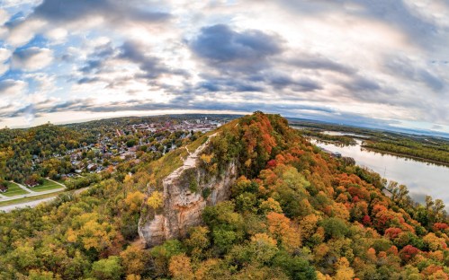 Aerial view of a rocky hill surrounded by vibrant autumn foliage and a river, overlooking a small town below.