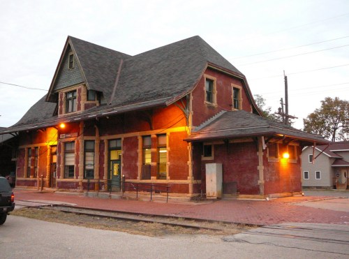 Historic brick train station with a steep roof, warm lights, and a surrounding paved area at dusk.