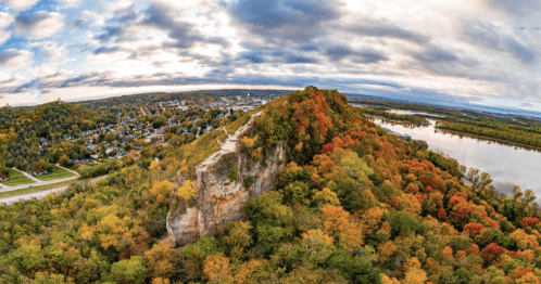 A panoramic view of a colorful autumn landscape with a rocky hill, river, and a town in the background under a cloudy sky.