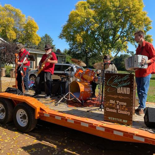 A band performs on a trailer stage, featuring musicians with instruments, surrounded by trees and a sunny sky.