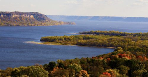 A scenic view of a river surrounded by lush greenery and autumn-colored trees under a clear blue sky.