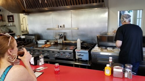 A woman drinks soda at a red counter while a cook prepares food at a grill in a diner kitchen.