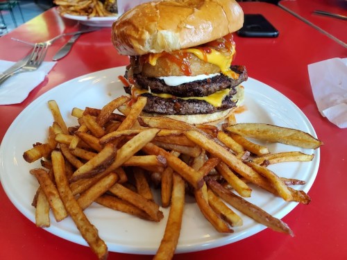 A towering burger with cheese, sauce, and onion rings, served with a side of golden French fries on a red table.
