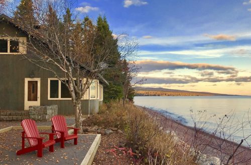 A serene lakeside scene featuring two red chairs, a cabin, and a calm water view under a colorful sky.