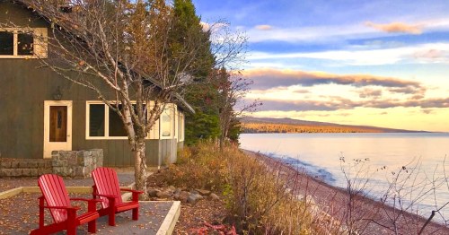 A cozy cabin by the shore with two red chairs, surrounded by trees and a calm lake under a colorful sunset sky.
