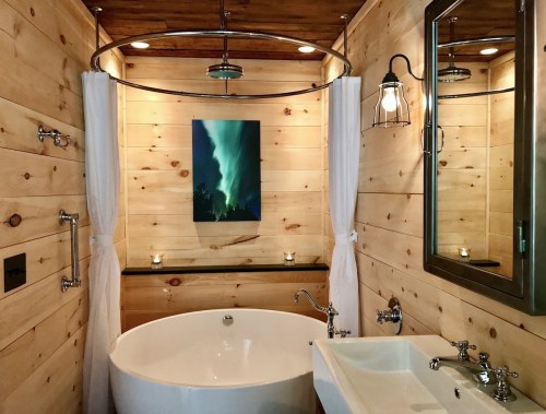 A cozy bathroom with wooden walls, a freestanding tub, a sink, and a painting of the northern lights. Soft lighting and candles.