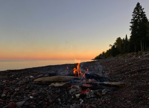 A campfire burns on a rocky beach at sunset, with trees silhouetted against a colorful sky.