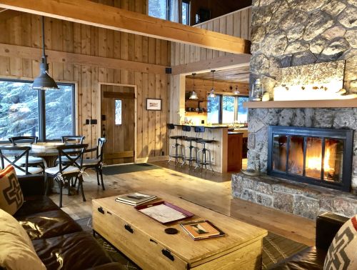 Cozy cabin interior with wooden walls, a stone fireplace, and a warm living area featuring a coffee table and dining space.