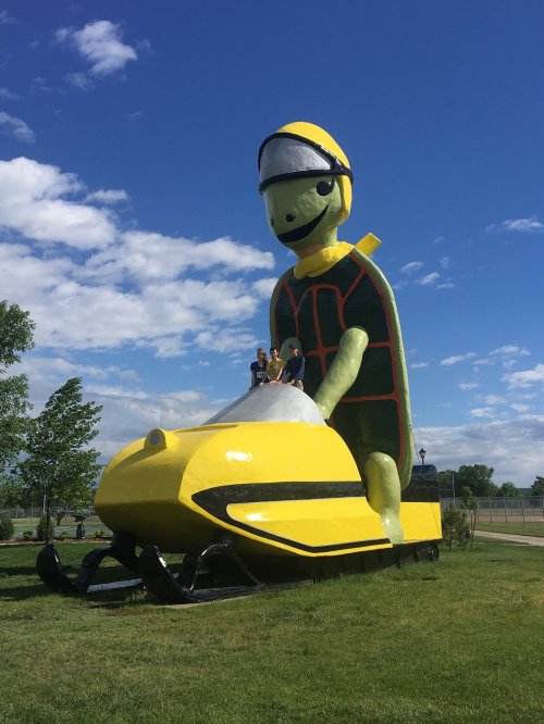 A large turtle statue wearing a helmet, with people sitting on a yellow snowmobile in a grassy area under a blue sky.