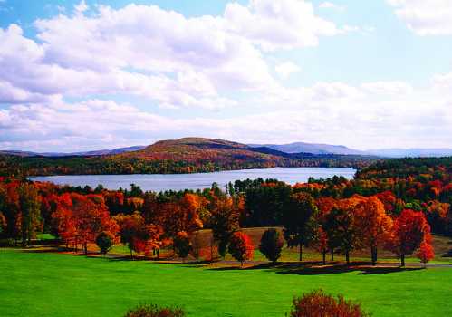 A scenic view of a lake surrounded by vibrant autumn foliage and rolling hills under a partly cloudy sky.