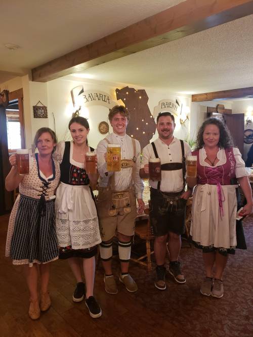 Five people in traditional Bavarian attire hold up large beer mugs, smiling in a cozy indoor setting.