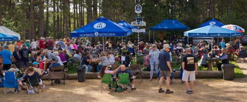 A large crowd gathers under blue tents at an outdoor event, enjoying food and music in a forested area.