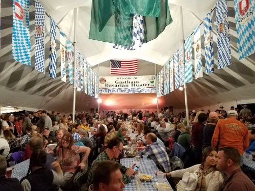 A lively indoor festival scene with people dining under a large tent decorated with banners and flags.