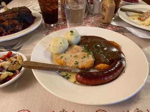 A plate of food featuring breaded meat, sausage, dumplings, and gravy, with a side salad and drinks in the background.