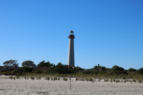 A tall lighthouse stands against a clear blue sky, surrounded by sandy beach and greenery.