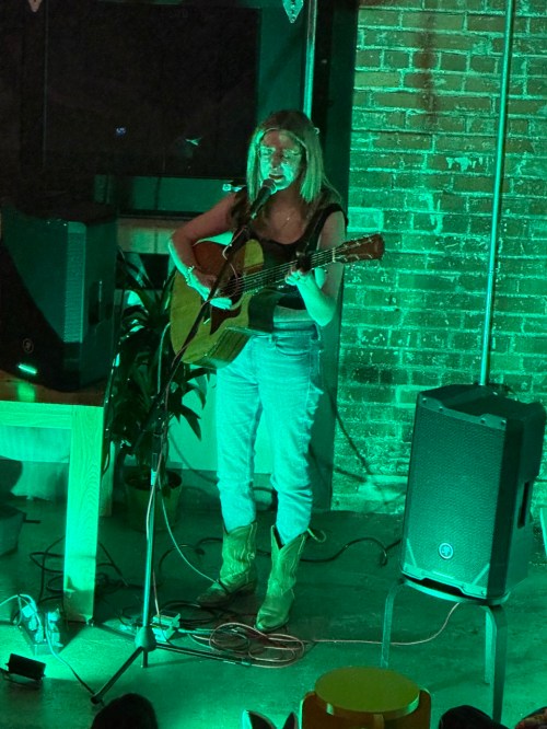 A musician performs on stage with an acoustic guitar, wearing cowboy boots, under green lighting in a cozy venue.