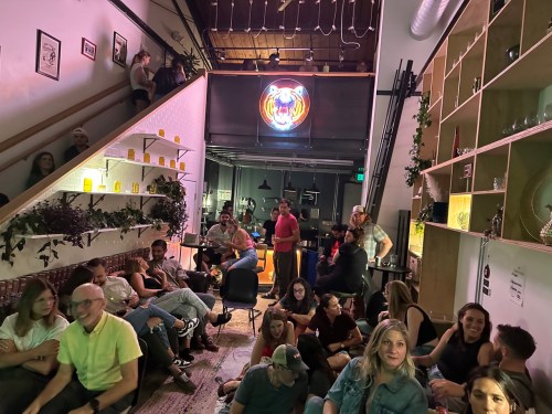 A lively indoor gathering with people sitting and standing, colorful decor, and a neon sign in the background.