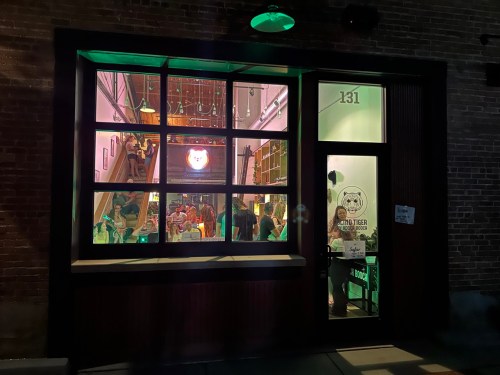 A brightly lit café with a large window, showcasing people inside and a neon sign, set in a brick building.