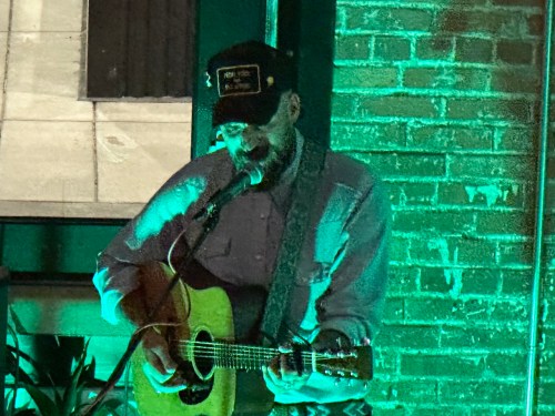 A musician performs with an acoustic guitar in a dimly lit venue, illuminated by green light.