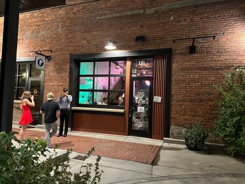 A group of people outside a brick building with colorful lights in the window, enjoying the evening atmosphere.